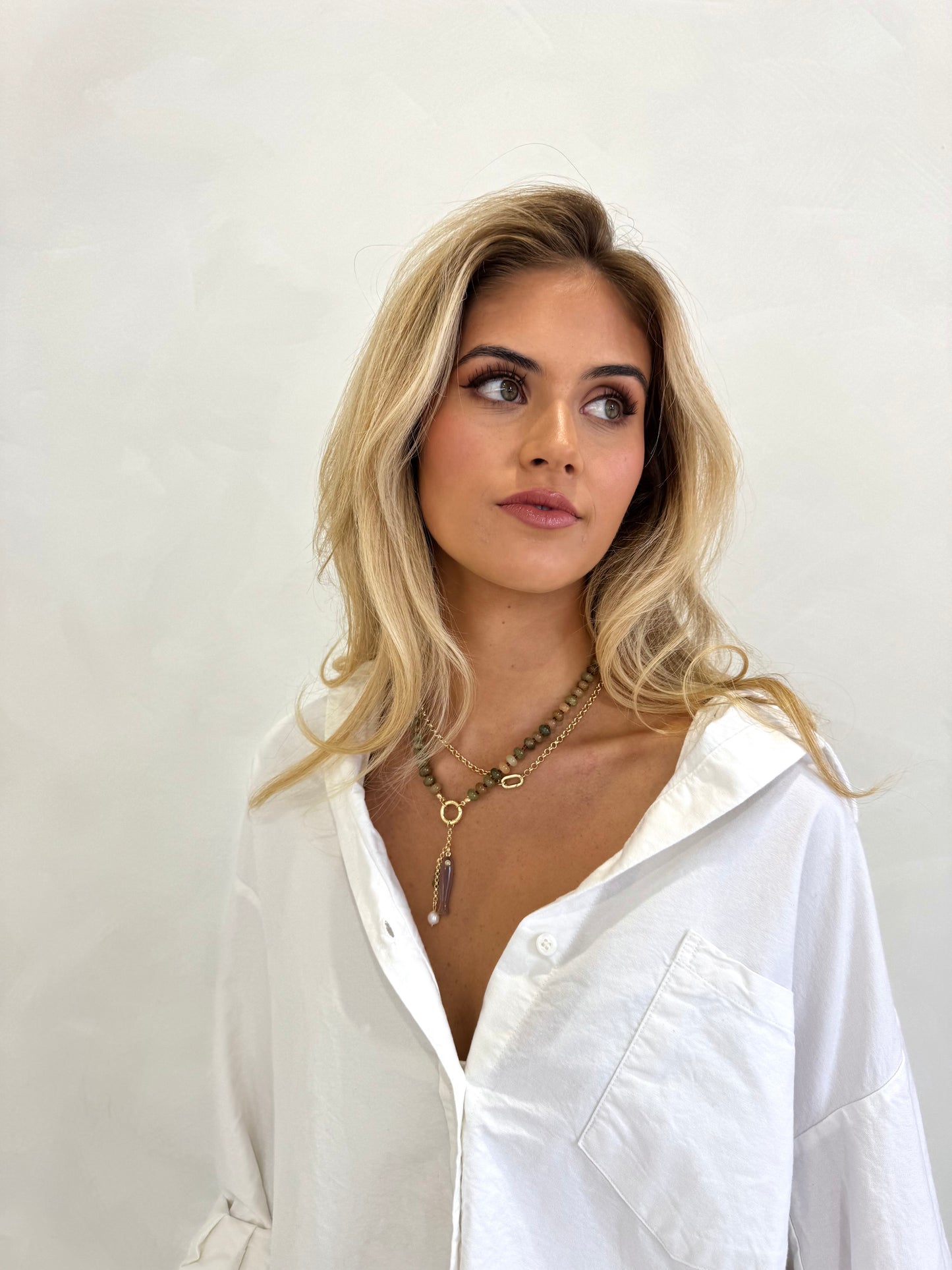 A blonde woman wearing a white button-down shirt and layered gold necklaces, posing against a plain off-white wall.