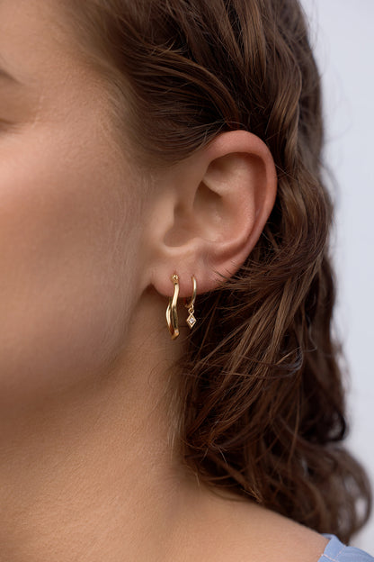 Close-up of a person wearing Lani Wave Hoops - Gold earrings with a blurred background