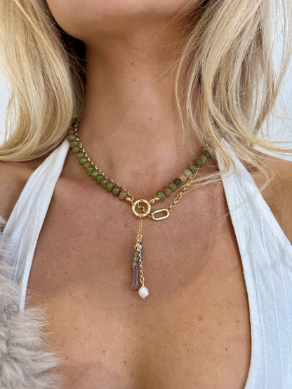 A close-up of a model wearing a bohemian-style gold chain necklace with green beads, a feather charm, and a white pearl drop, paired with a white halter top.